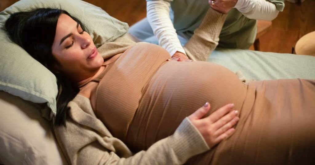 A pregnant woman lying on a bed and breathing out through the mouth as her midwife holds her hand and feels her belly.