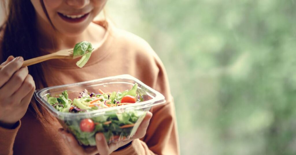 A woman holds a wooden fork and prepares to take a bite of salad from a clear container. There's a green blurred background.