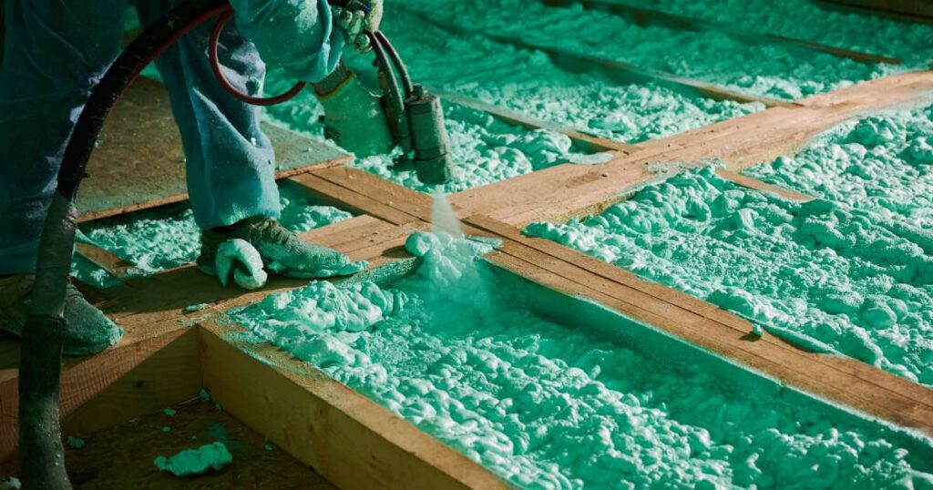 A man hunching over to insulate the wooden framing of a home's floor with green spray foam insulation.