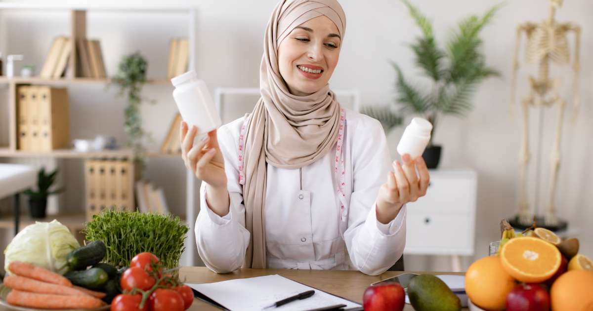 A professional nutritionist sitting at a table holding two pill bottles. Different foods surround her.