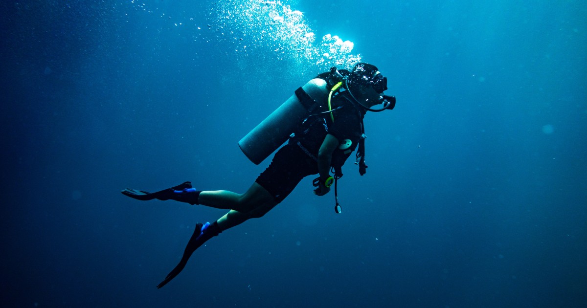 A person in a full wetsuit with scuba diving gear on swimming upward toward the light in a dark blue ocean.