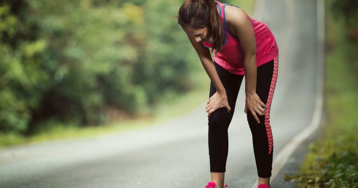 A woman in workout gear stands on the side of a running path. She leans forwards with her hands braced on her knees.