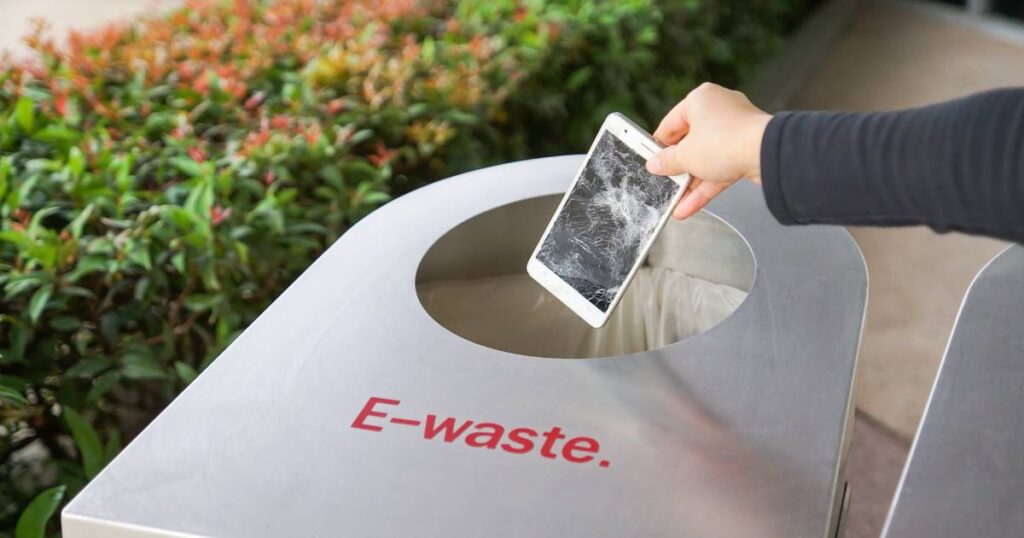 A person's hand drops a damaged smart phone with a shattered screen into a container marked E-waste.