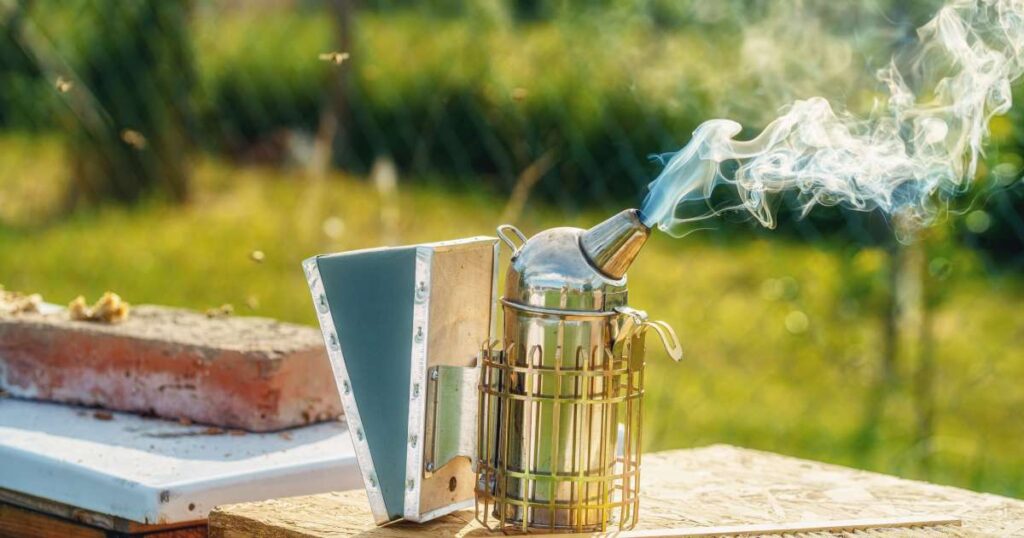 A silver beekeeping smoker sits on top of a wooden hive box. Another wooden hive box sits behind it.