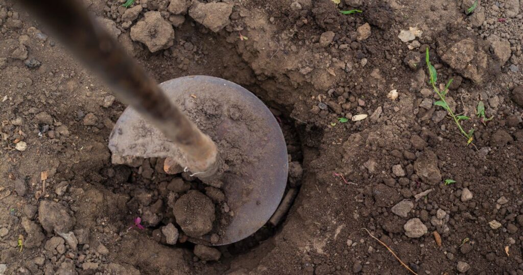 A farmer using a large auger attached to a ground drill drilling holes for plants or posts on farmland.