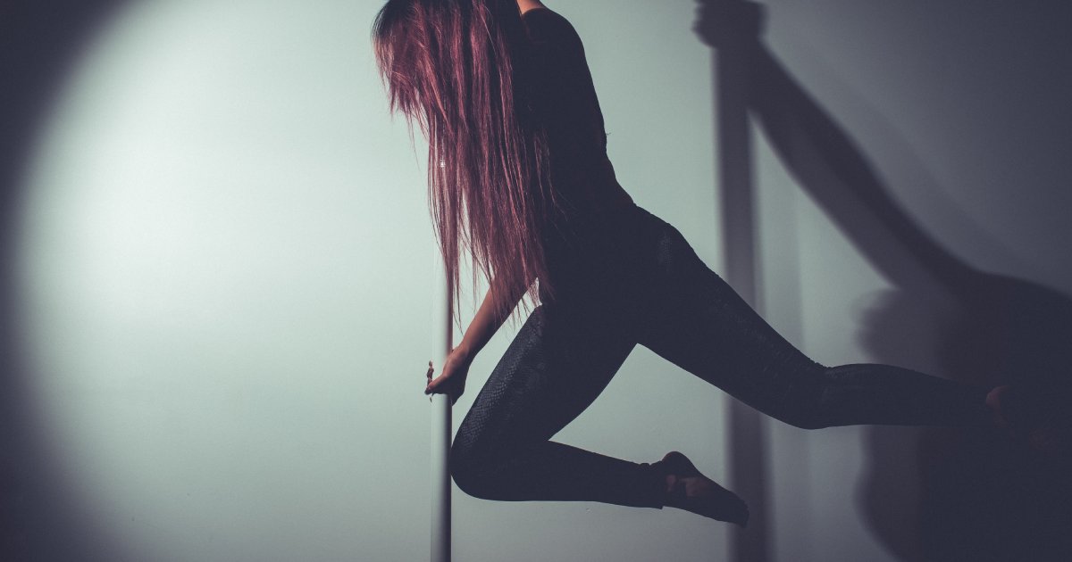 A light shines on a person gripping an aerial pole. She hangs in the air with her knees flexed and her feet behind her.