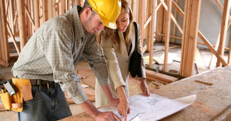 A contractor with a homeowner standing at a table inside a new build. They're discussing a blueprint for the home.