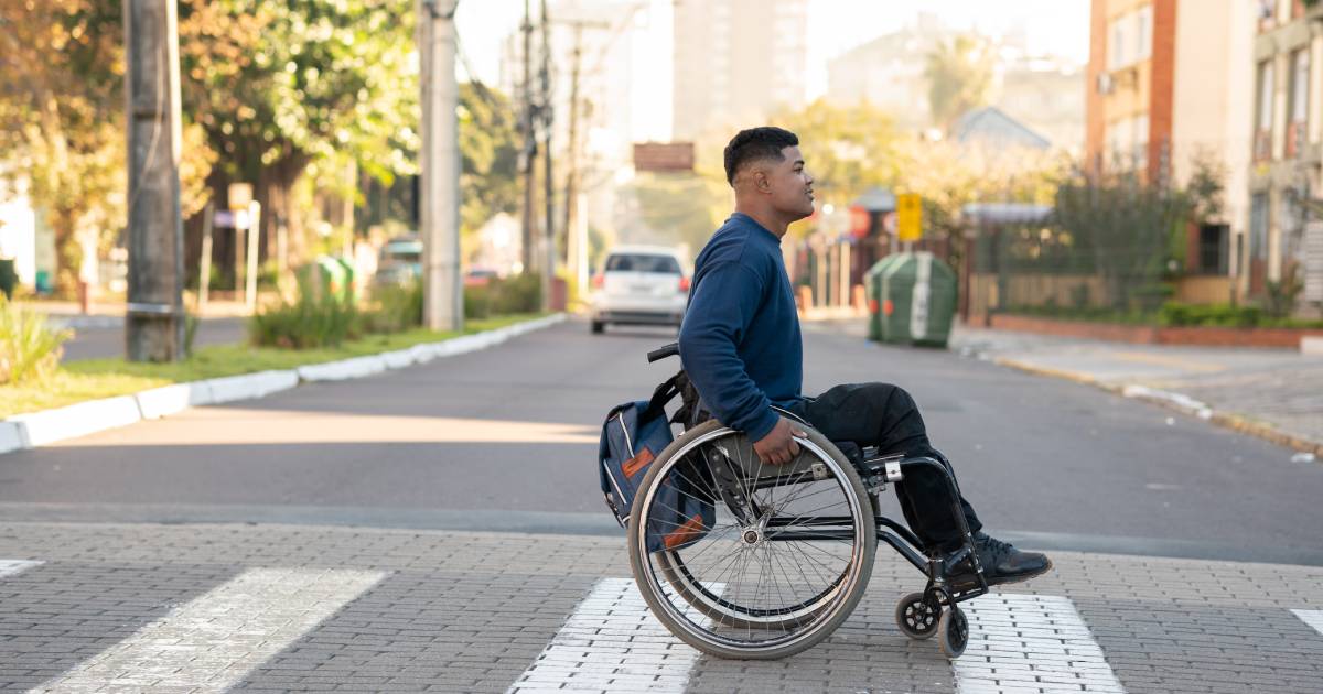 A man in a wheelchair crosses a street. He has a backpack on his chair, and there is a white car in the background.