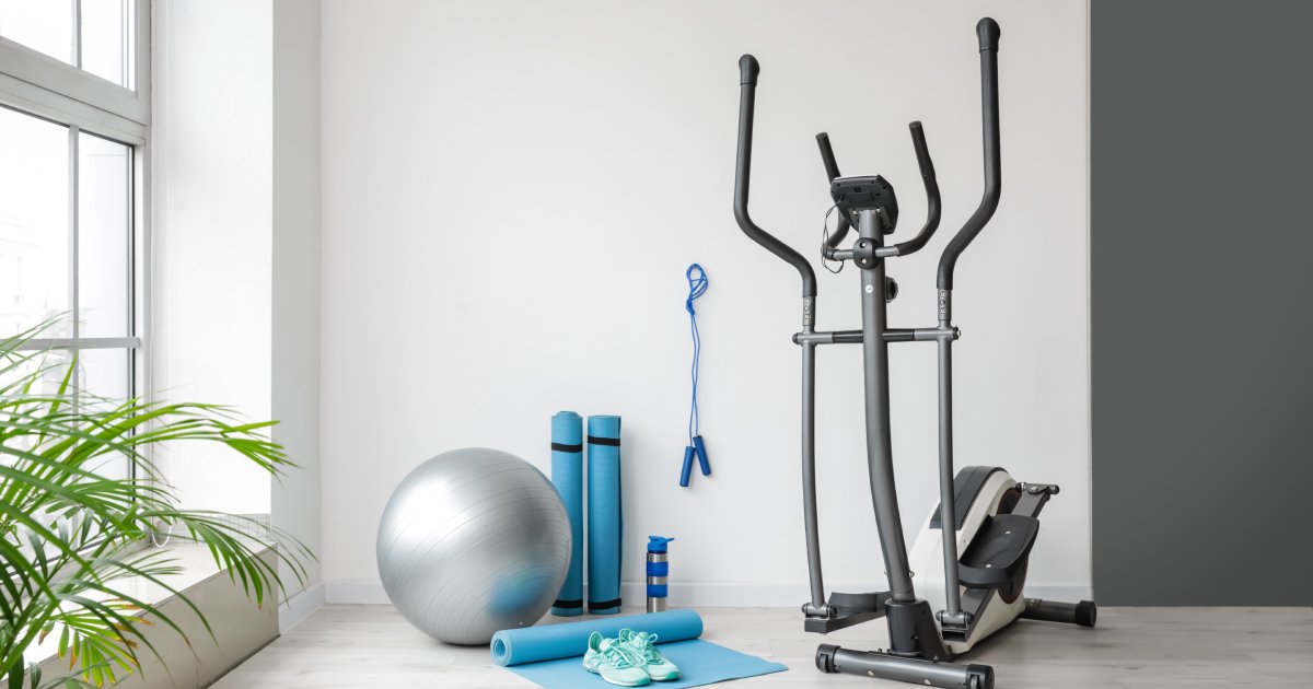 A bright room with fitness equipment, including three blue yoga mats, a silver exercise ball, a jump rope, and an elliptical.