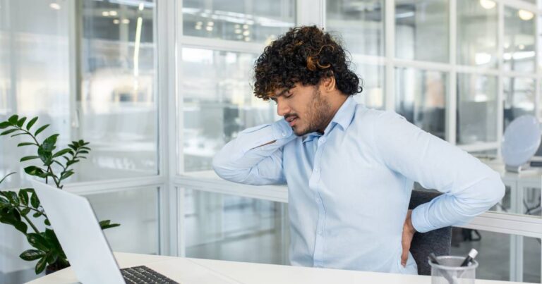 Male employee wearing a blue button-down shirt holding his neck and back while sitting behind an office desk with a laptop.