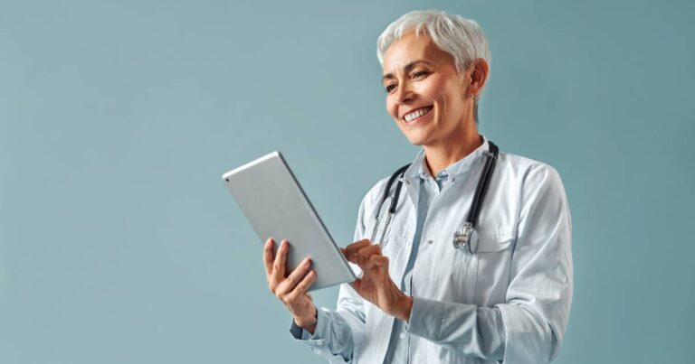 A female healthcare professional stands against a mint green background, holding a tablet and a stethoscope around her neck.