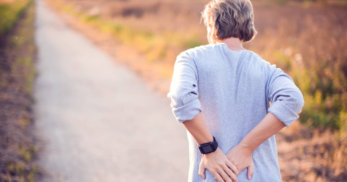 A woman wearing a black smartwatch and gray shirt while taking a walk on an outdoor path. She is holding her lower back.